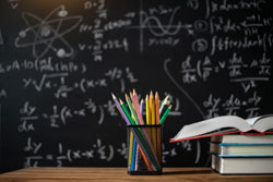 Several pencils in a pencil holder next to books on a table in front of a blackboard containing chalk notes on physics.